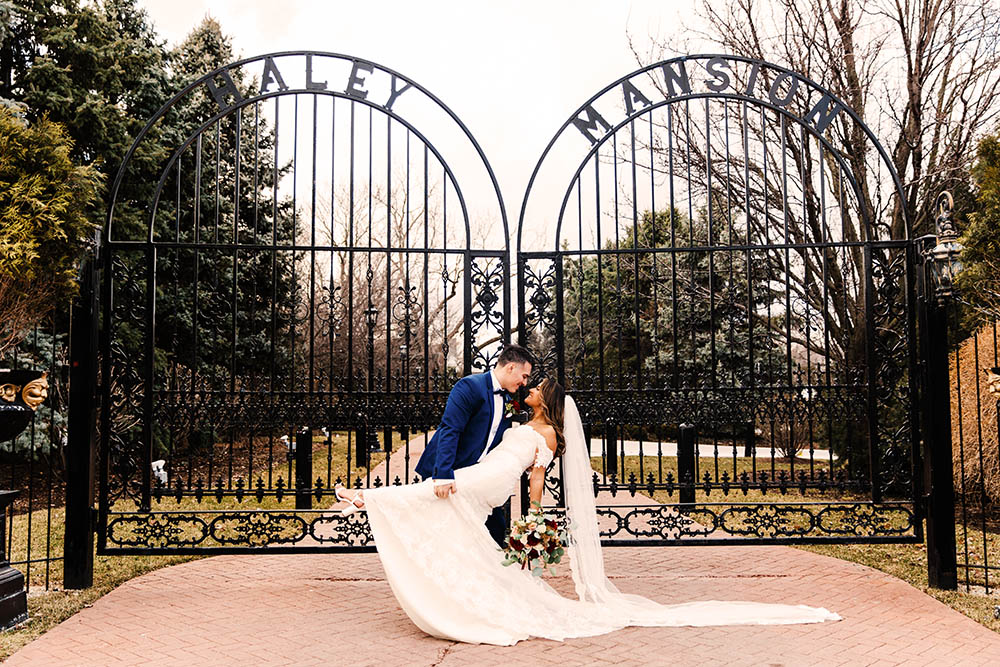 A couple in wedding attire poses romantically in front of an ornate black wrought-iron gate with Haley Mansion written at the top. The groom in a blue suit dips the bride, who holds a bouquet, on a brick pathway. Trees fill the background.