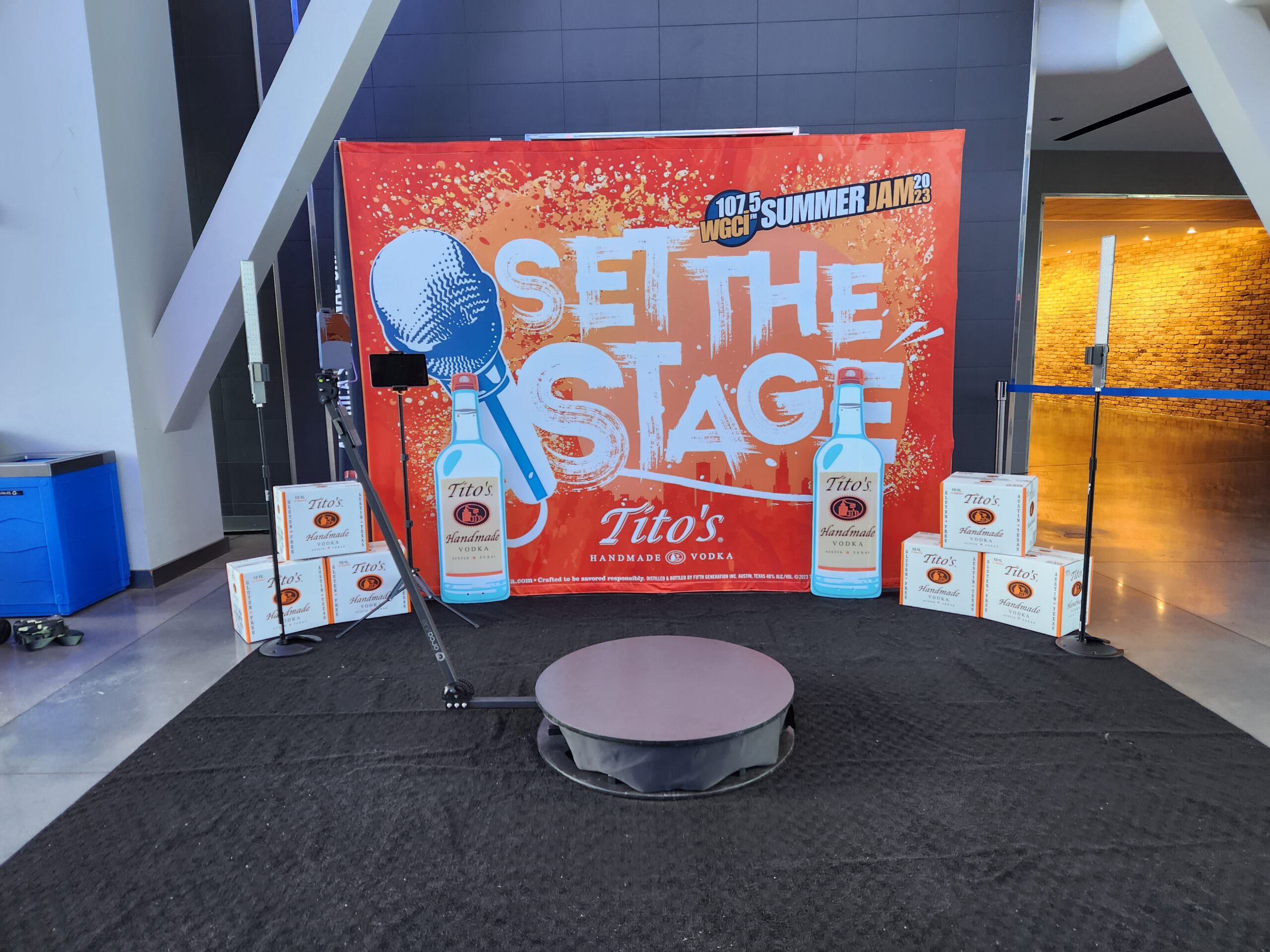 A stage setup featuring a banner with Set the Stage and the Titos Handmade Vodka logo. Several boxes of Titos are arranged around the stage. A microphone stand and a circular platform are positioned in front.