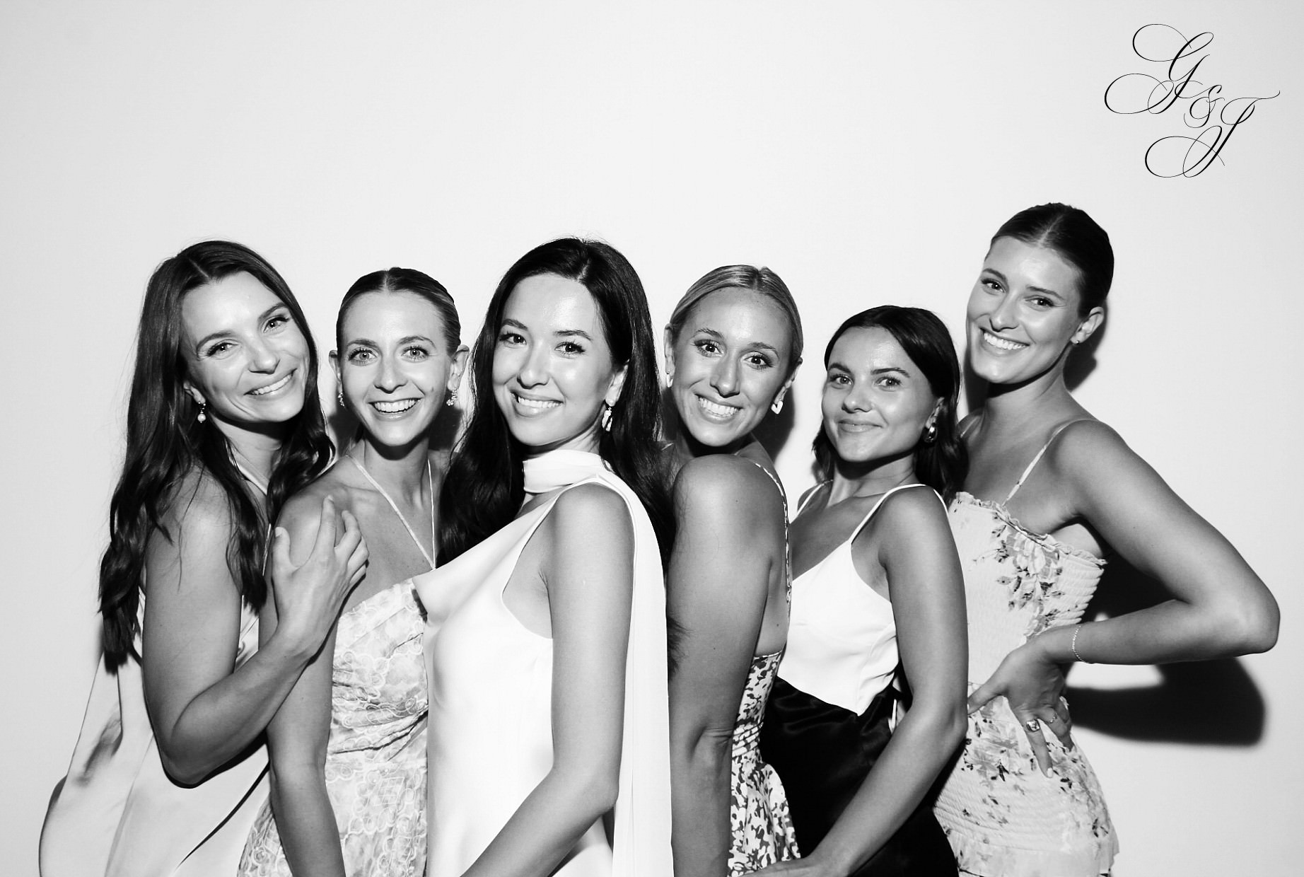 Six smiling women in elegant dresses stand closely together in front of a white background. The photo is in black and white, and theres a monogram in the top corner.