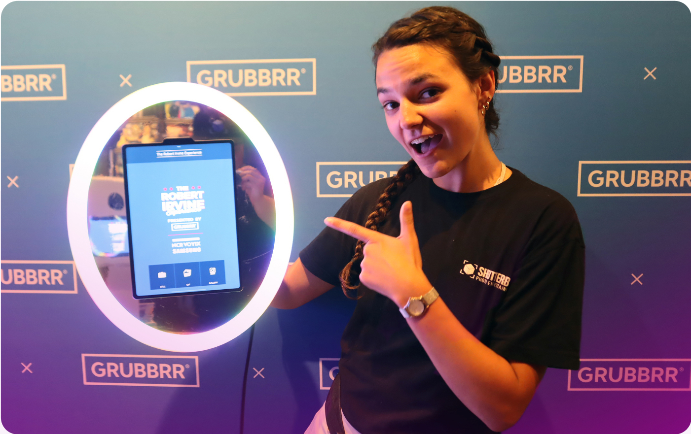 A smiling woman points at a circular kiosk display with a touchscreen, reminiscent of a roaming photo booth. The background boasts a blue and purple gradient with the word GRUBBRR repeated across it. Sporting a black t-shirt with a logo, they capture curiosity and innovation all at once.