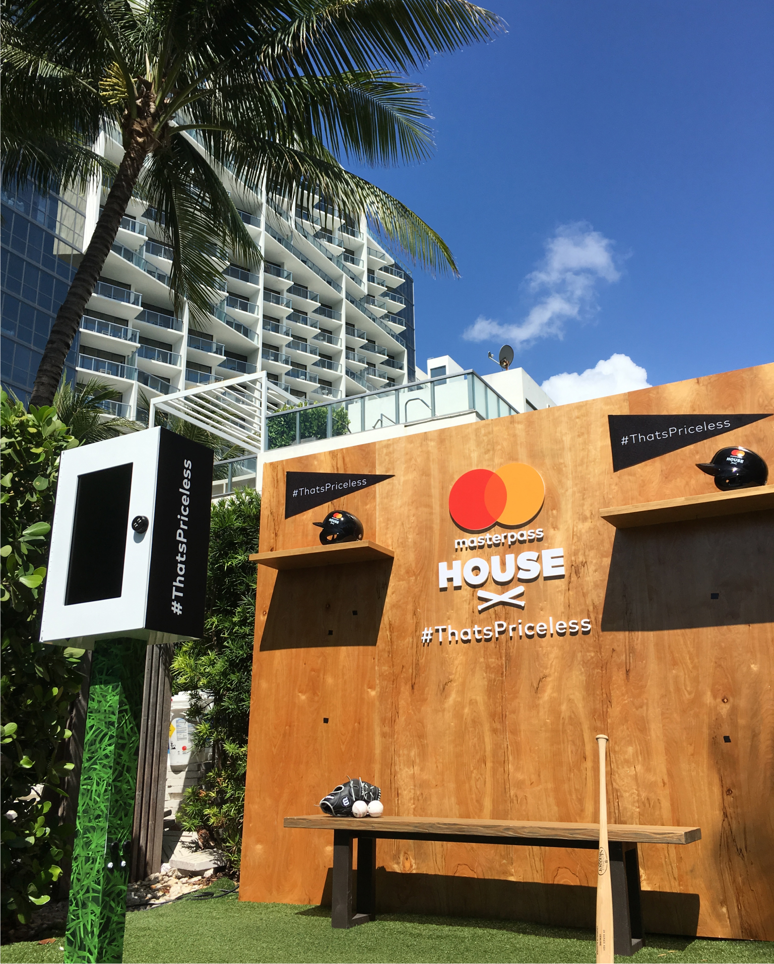 Outdoor promotional setup for Masterpass House with a wooden backdrop displaying logos and the hashtag #ThatsPriceless. Baseball gear like a bat and helmet are placed on a bench. Photography services for activations capture tall buildings and a palm tree in the background under a clear blue sky.