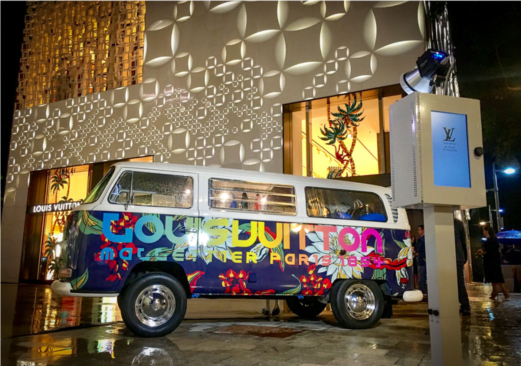 A colorful van with vibrant floral designs and the words Louis Vuitton and Neverfuller Paradise is parked in front of a lit-up luxury storefront, offering an immersive company-experiential-service. A nearby kiosk displays the Louis Vuitton logo amidst a nighttime city setting.