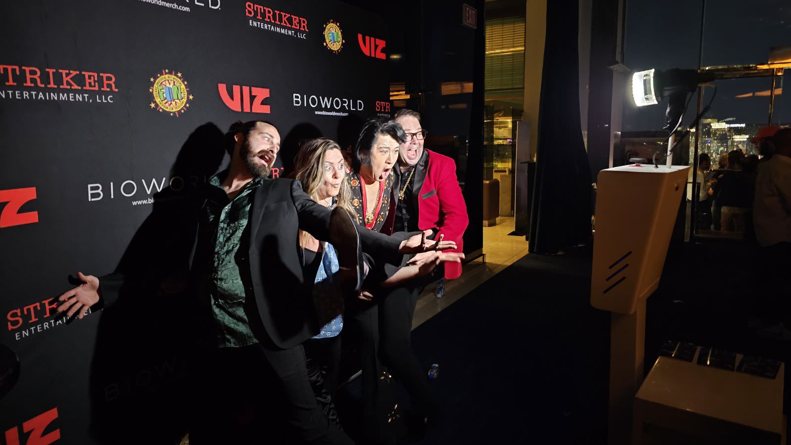 Four people striking a playful pose on a red carpet at the Las Vegas event, with a backdrop featuring logos from Striker Entertainment, Bioworld, and Viz. They appear excited, as if taking a group selfie in the lively photo booth setting.