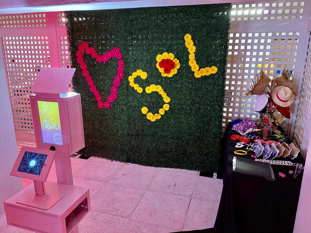 A Milwaukee Photo Booth Rental features a pink kiosk and a lush green foliage backdrop adorned with colorful flowers spelling Sol and a heart. To the right, a table offers props like hats and glasses for photo sessions, adding fun to any event.