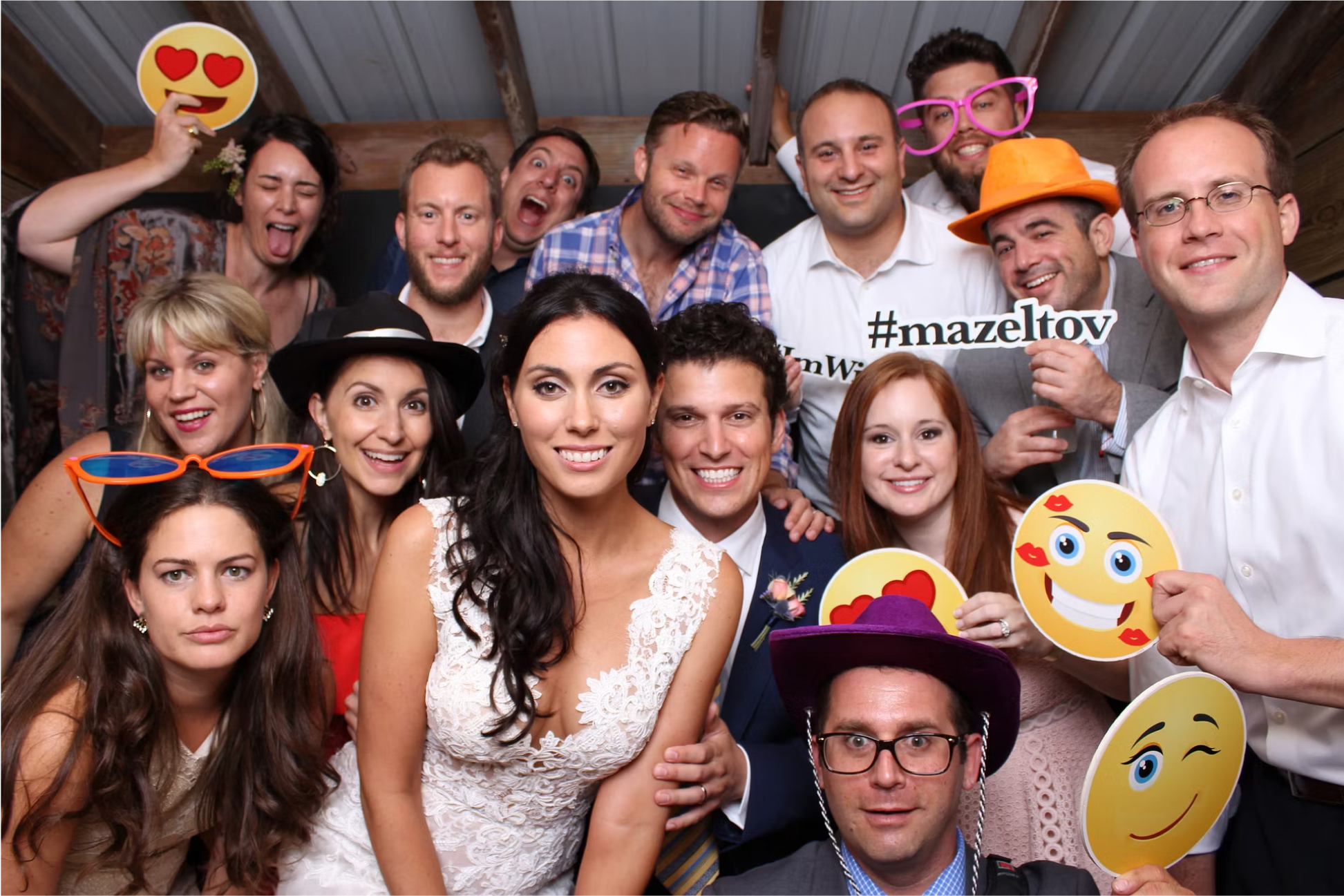 A group of people at a festive event, posing for a fun photo from wedding photo booth rentals. They are wearing colorful hats and glasses, holding emoji props and a #mazeltov sign. Everyone is smiling or making playful expressions.