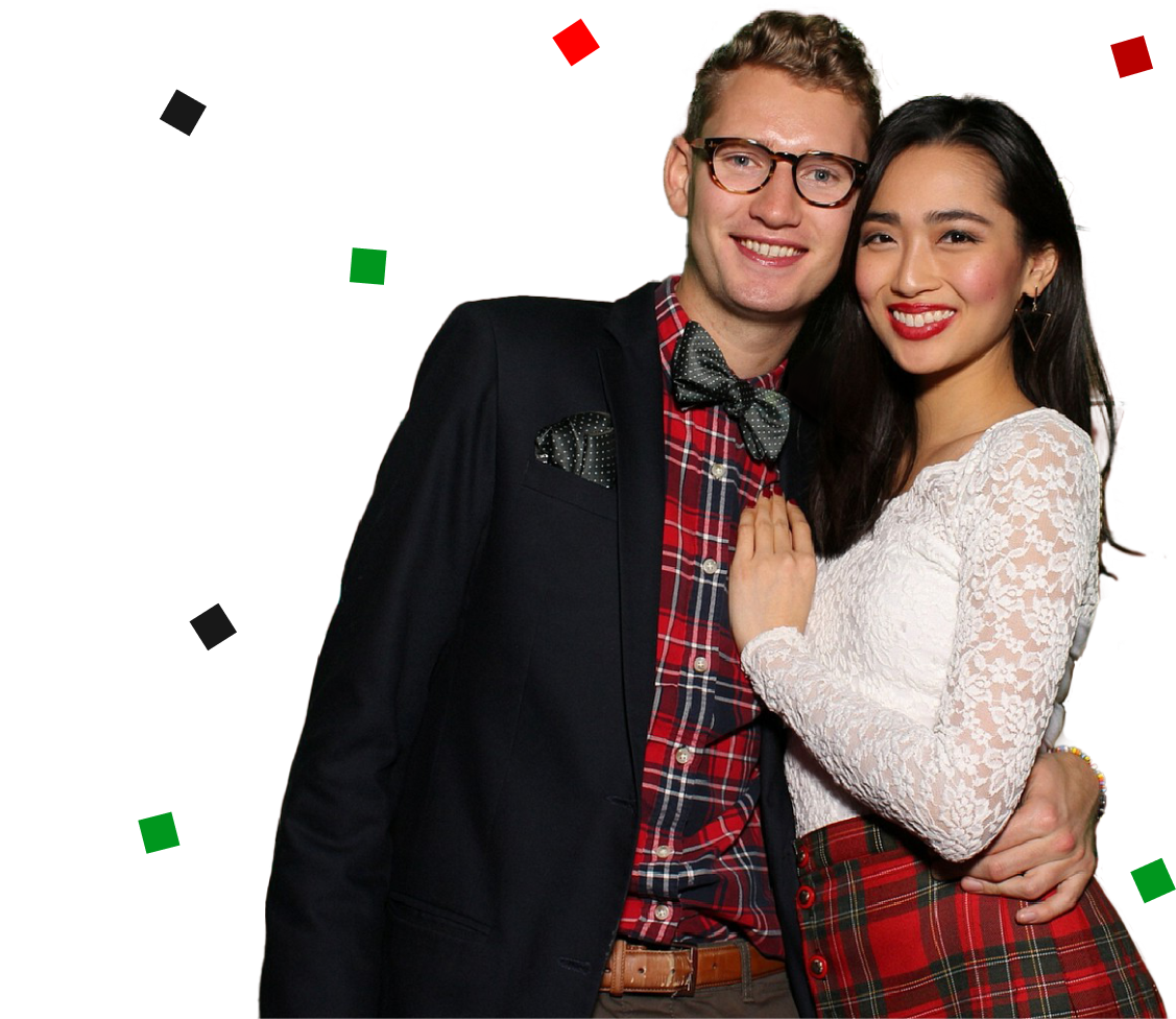 A man in a suit, glasses, and bow tie warmly embraces a woman in a lace top and tartan skirt. They smile amidst scattered colored squares—a festive moment perfect for holiday parties captured by the Photo Booth Rental Company.