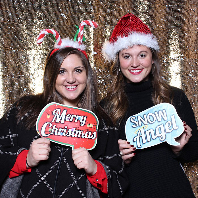 Two women smiling in front of a glittery backdrop, holding festive signs saying Merry Christmas and Snow Angel. They are wearing holiday-themed accessories: one with candy cane antlers and the other with a Santa hat, capturing the joyful spirit at a Christmas party photo booth.