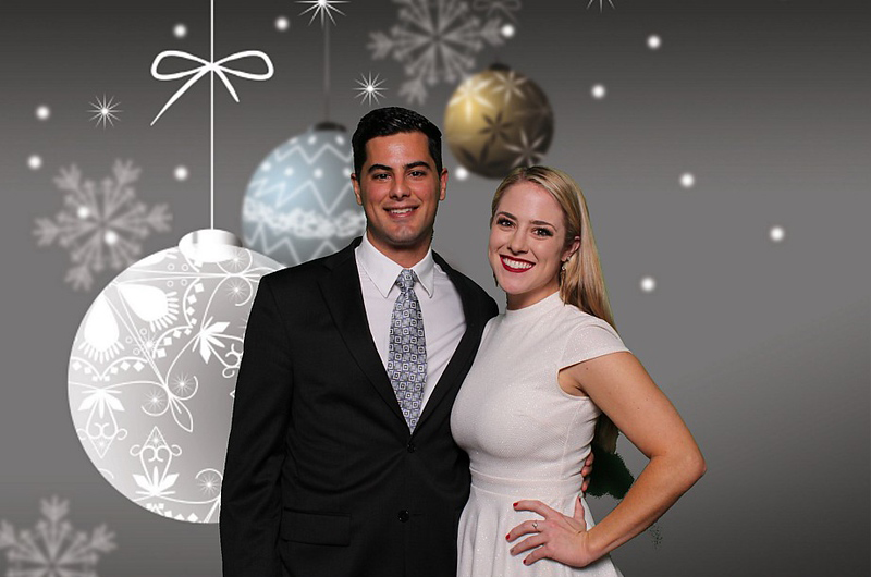 A man in a suit and a woman in a white dress pose together, smiling in front of a festive holiday background with hanging ornaments and snowflakes, capturing the joy of the Christmas photo booth.