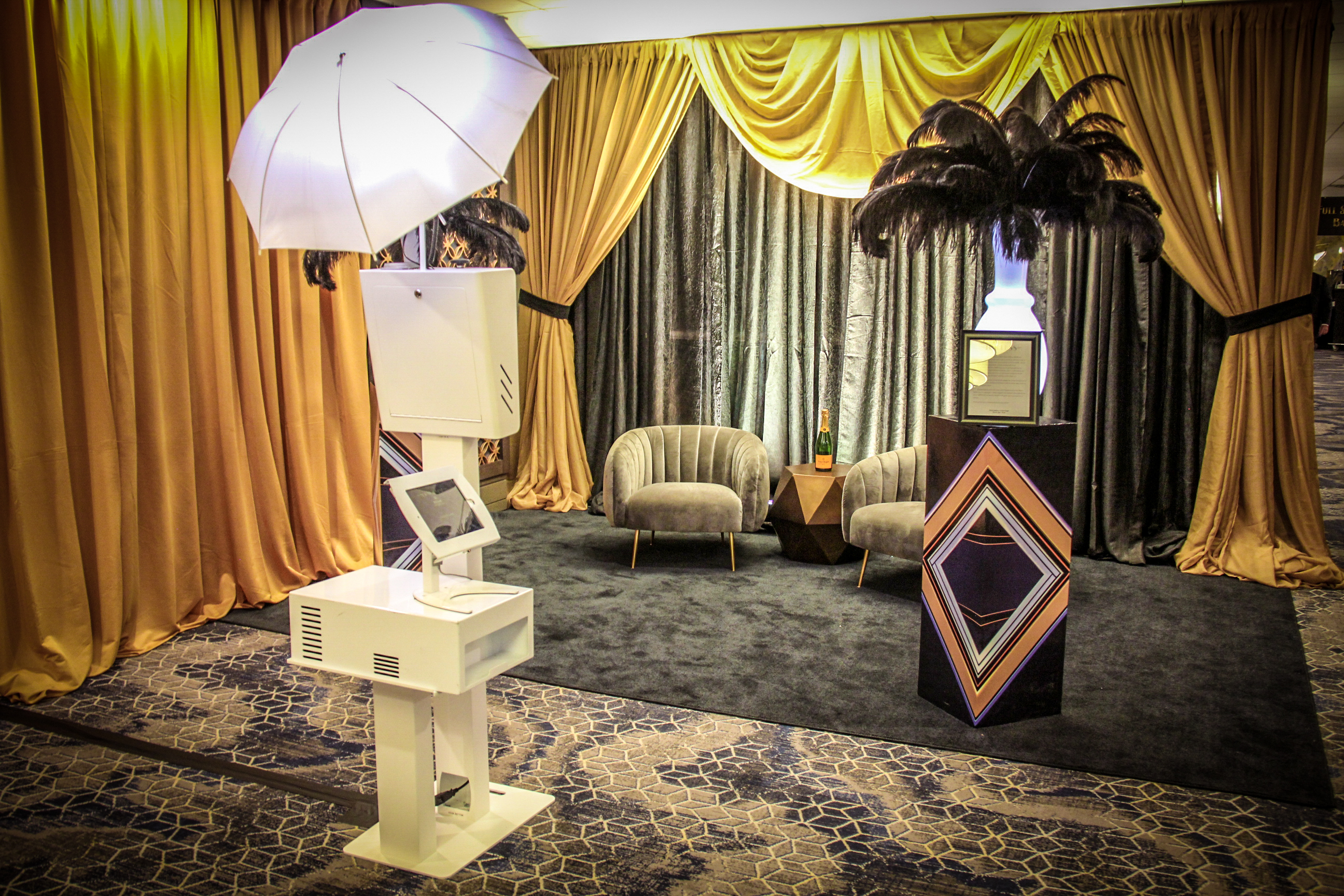 A stylish holiday party setup features a sleek ShutterBox photo booth in a room with gold and gray drapes. Two gray chairs flank a small brown table on a dark rug. An ornate stand holds feathered decor, while the modern booth with its white umbrella awaits guests.