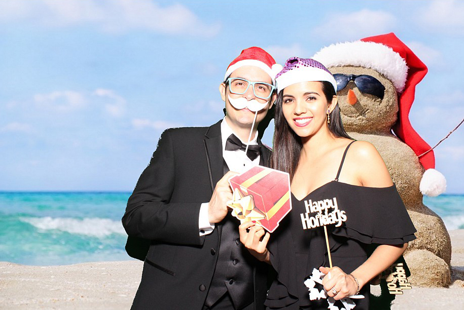 A man and a woman pose on a Miami beach, turning it into a festive photo booth scene with holiday party vibes. Sporting a Santa hat and reindeer antlers, they hold a gift box and a Happy Holidays sign, while a sand snowman with its own Santa hat completes the cheerful backdrop.