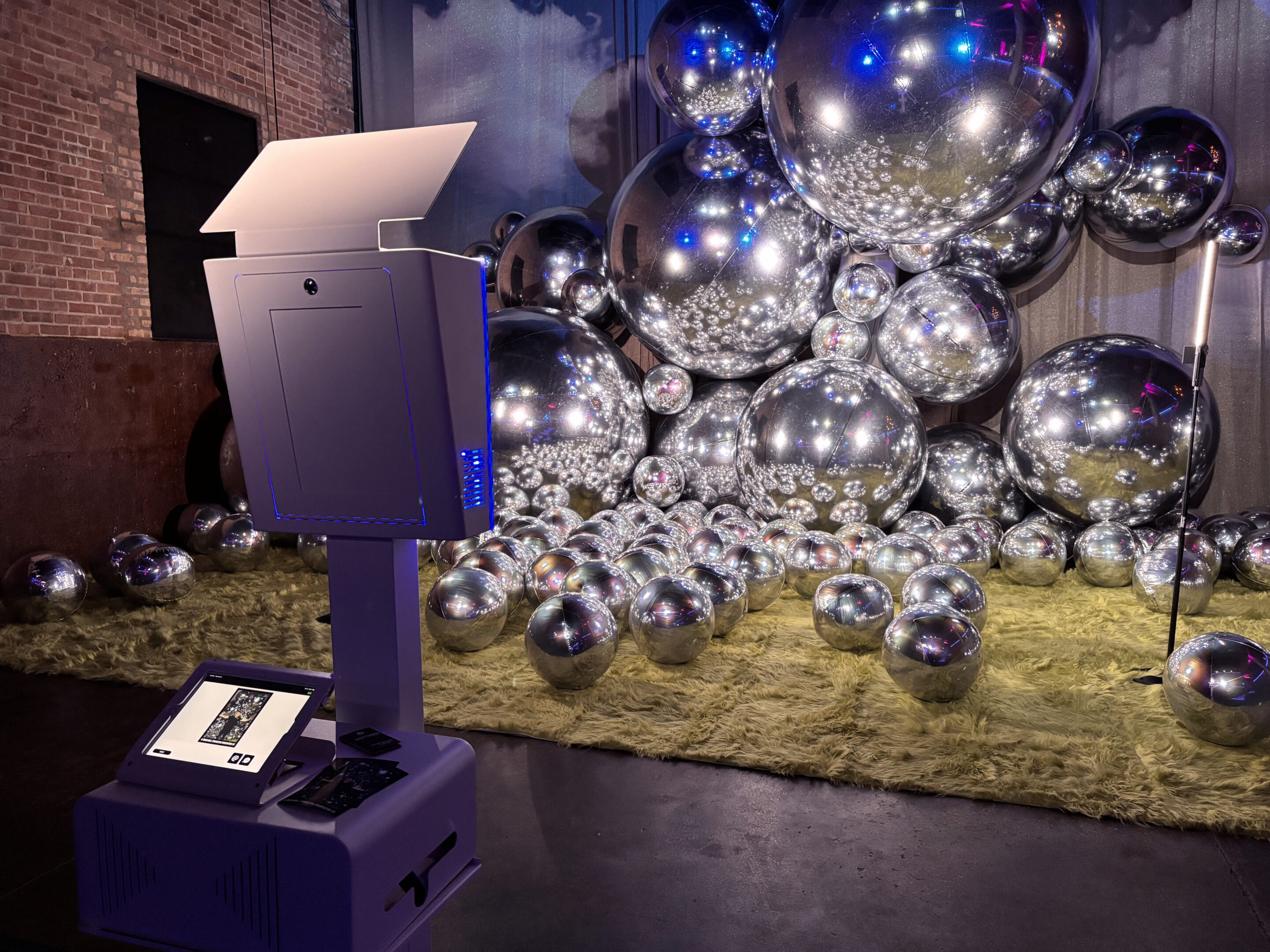 A modern photo booth rental in Chicago features a lit screen in a dimly lit room. Behind it, an artistic display of reflective silver spheres is elegantly arranged against a textured backdrop on a carpeted floor.