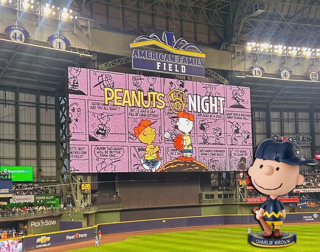 A large scoreboard at American Family Field displays Peanuts Night with comic strips; in the foreground, a Charlie Brown bobblehead poses like he’s ready for a Chicago Photo Booth Rental.