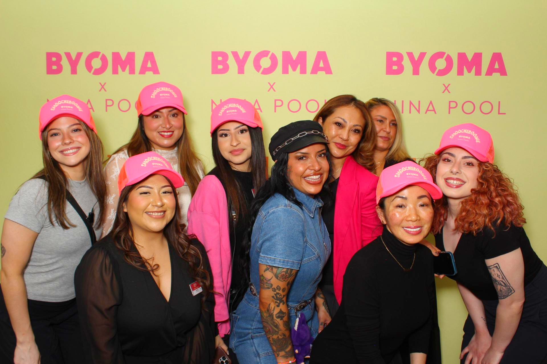 A group of nine women pose in front of a yellow BYOMA x NINA POOL backdrop, some in pink hats, at the Nina Pool Tiktok Star Meet & Greet in Chicago.