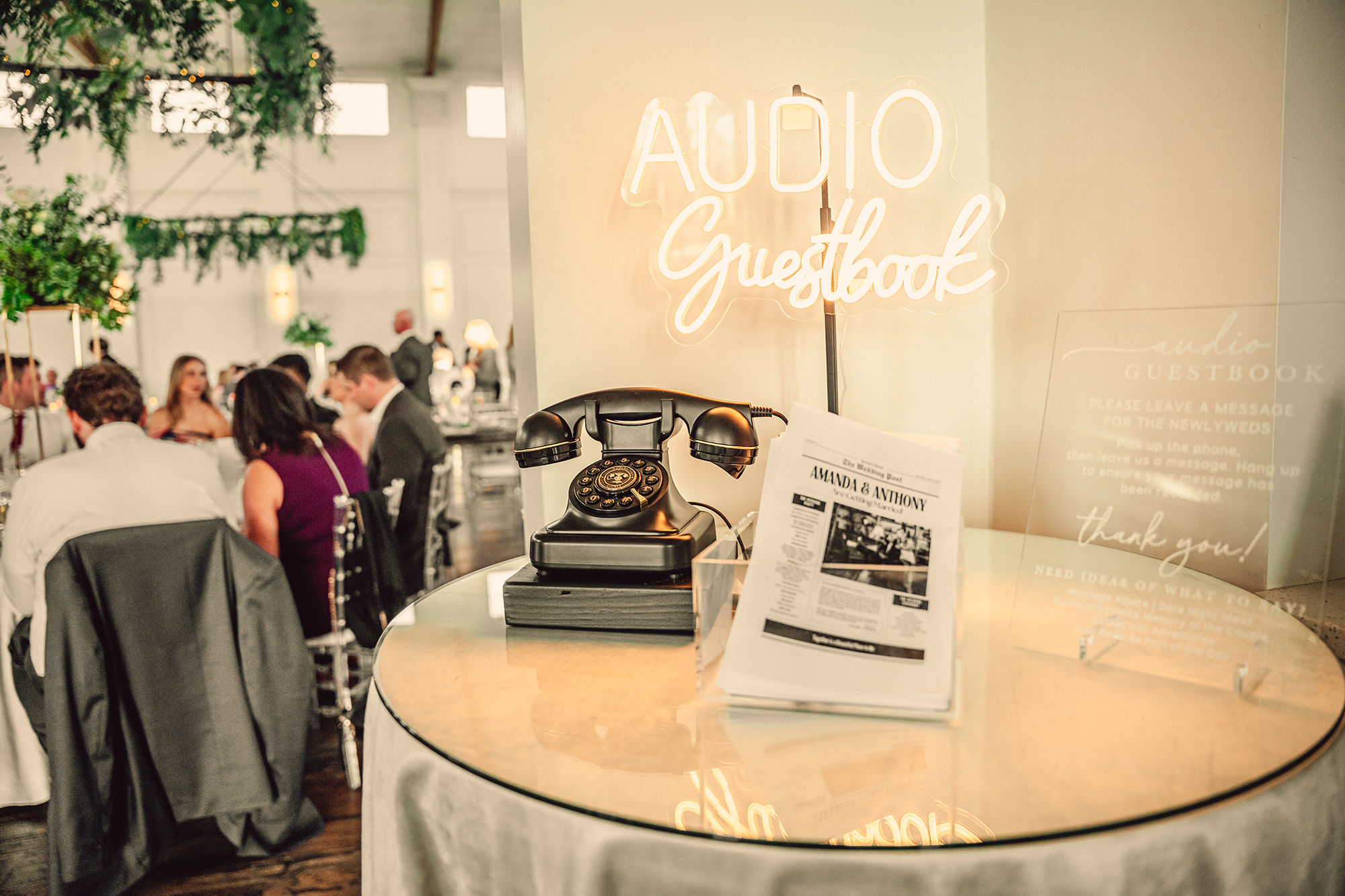 A vintage telephone sits on a round table beneath a neon Audio Guestbook sign at a wedding reception, adding charm alongside the buzz of guests and a nearby photo booth rental Chicago.