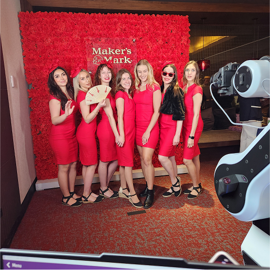 Seven women in matching red dresses pose in front of a red flower wall with a Makers Mark sign. A robotic camera arm is visible in the foreground taking their photo.