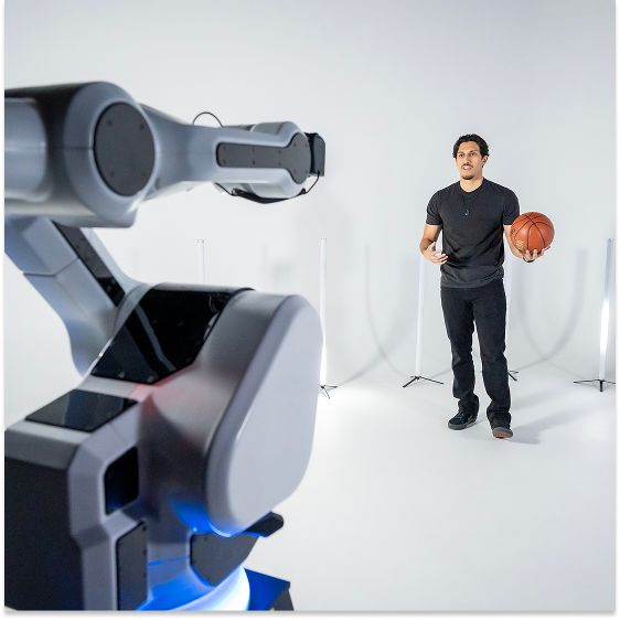 A man holding a basketball stands in front of a large robotic arm in a well-lit, minimalist studio setting.