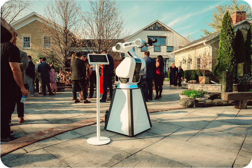 A robotic arm on a white stand is displayed outdoors near a touchscreen kiosk, with a group of people gathered in the background.