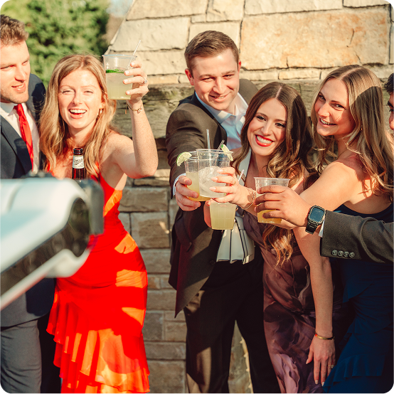 A group of five adults dressed in formal attire raise their drinks and smile for a photo outdoors in sunny weather.