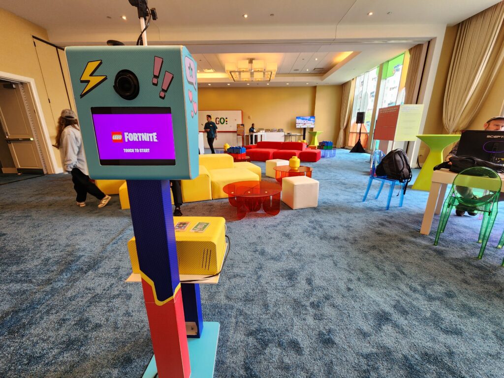 A Fortnite-themed kiosk stands in a colorful lounge with scattered seating, a TV screen, and guests—perfect to rent a photo booth in Chicago for your next gaming event.