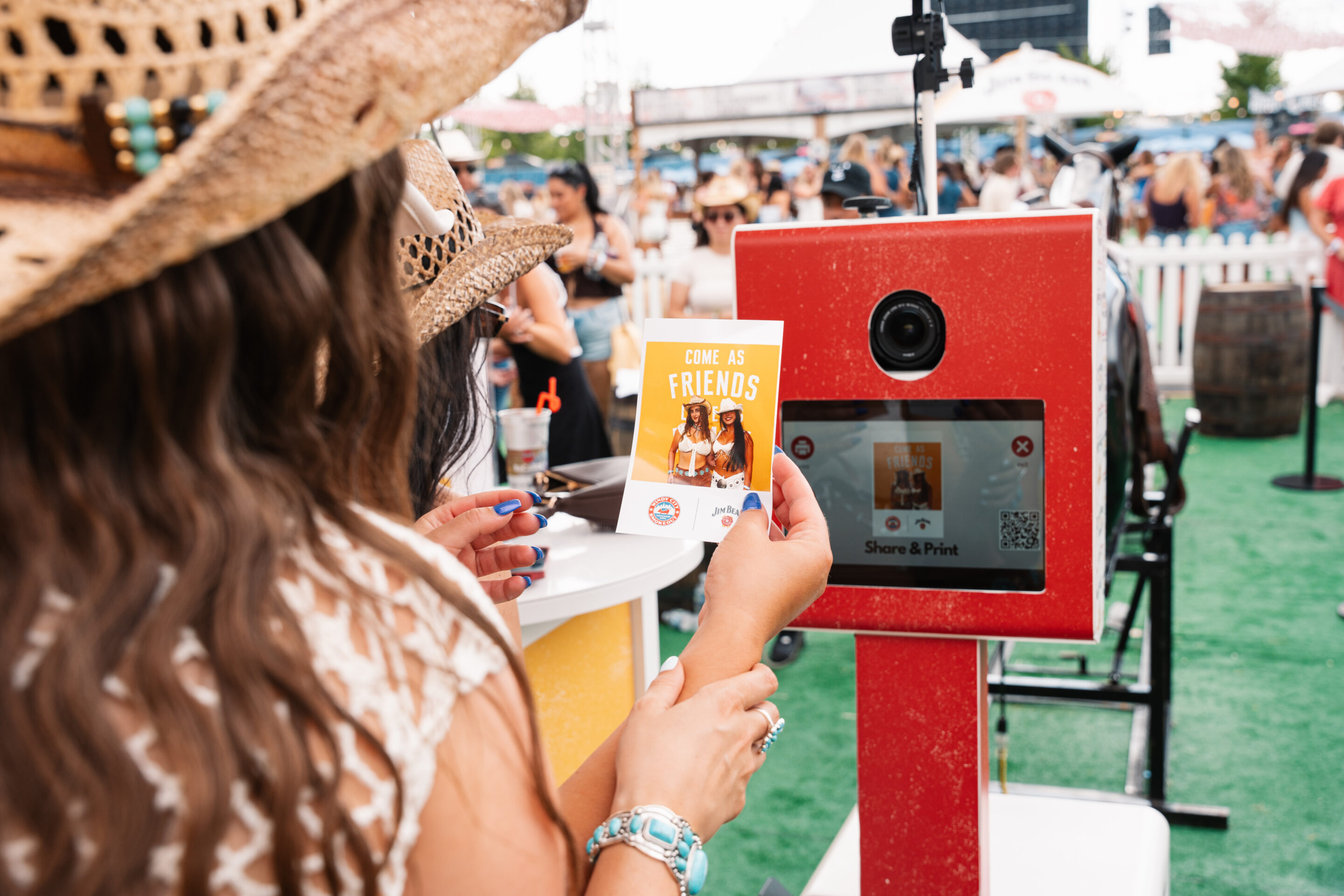 A person in a straw hat holds a printed photo labeled COME AS FRIENDS by a red Nationwide Photo Booth Rental at an outdoor event with people in the background.