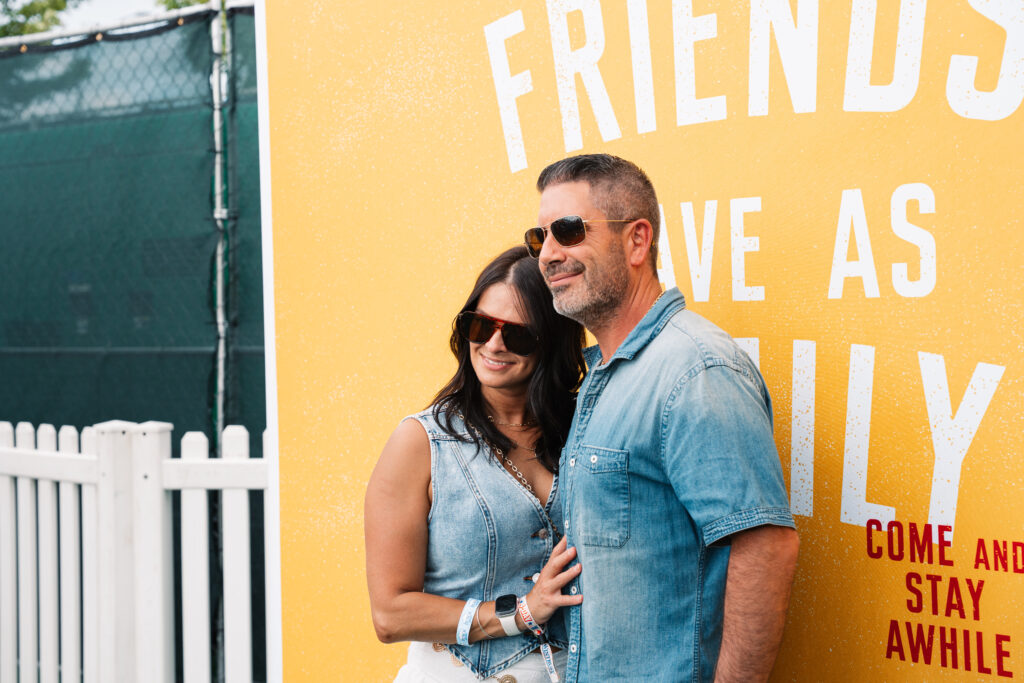 A woman and man wearing sunglasses and denim pose in front of a yellow wall, capturing a fun game day photo booth moment with bold white and red text as their backdrop.