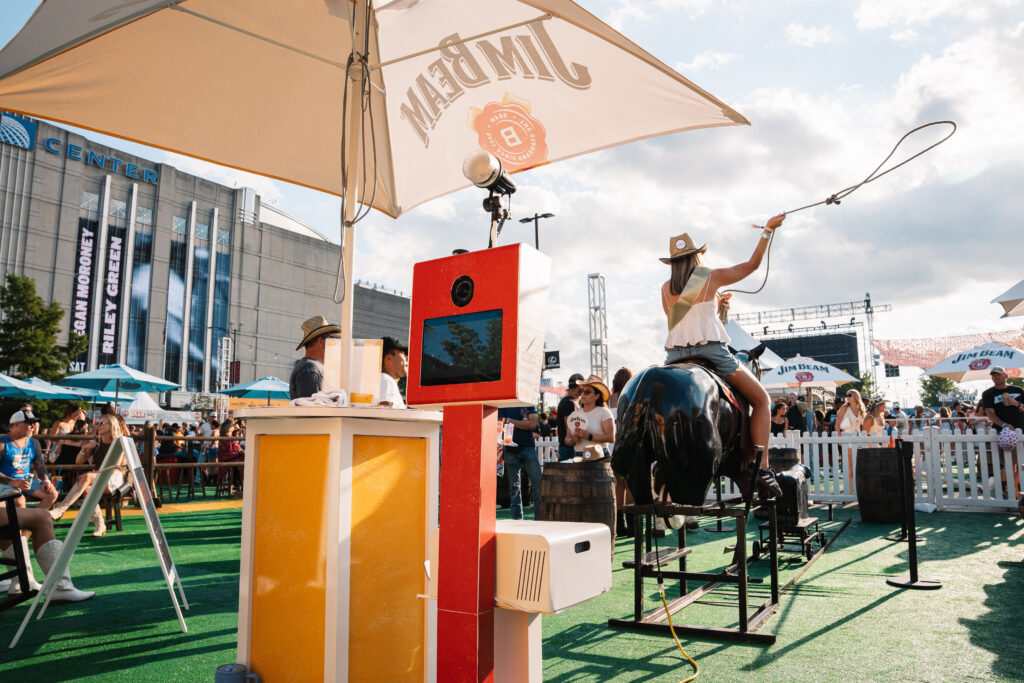 A woman rides a mechanical bull under a large branded umbrella at an outdoor event, near festival booths and a Nationwide Photo Booth Rental setup.