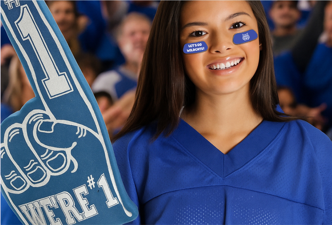 Young woman in a blue jersey holding a large foam finger that says WERE #1, with LETS GO WILDCATS! sticker on her cheek, surrounded by a crowd.