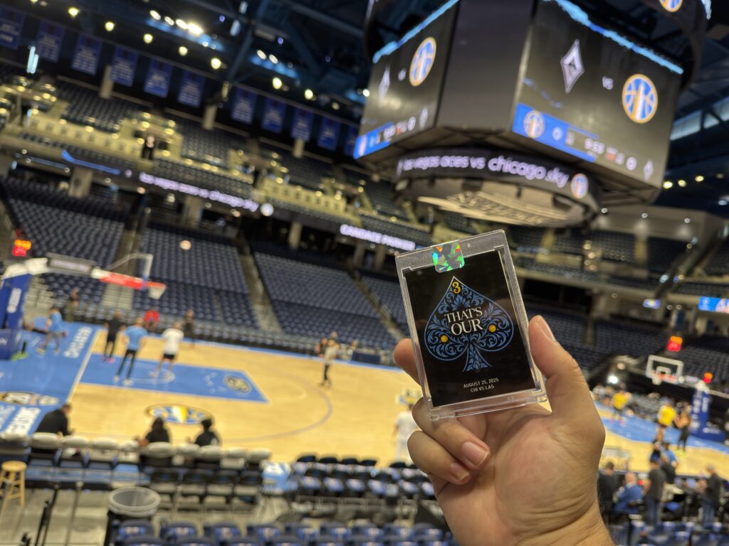 A hand holds a playing card encased in plastic at a basketball arena, where Brand Activation Photo Booth setups and team logos light up the big screens as players practice on court.