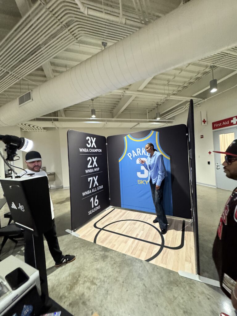 A man poses for a photo next to a large replica of a Chicago Sky jersey at an indoor brand activation photo booth with a wall listing basketball achievements.