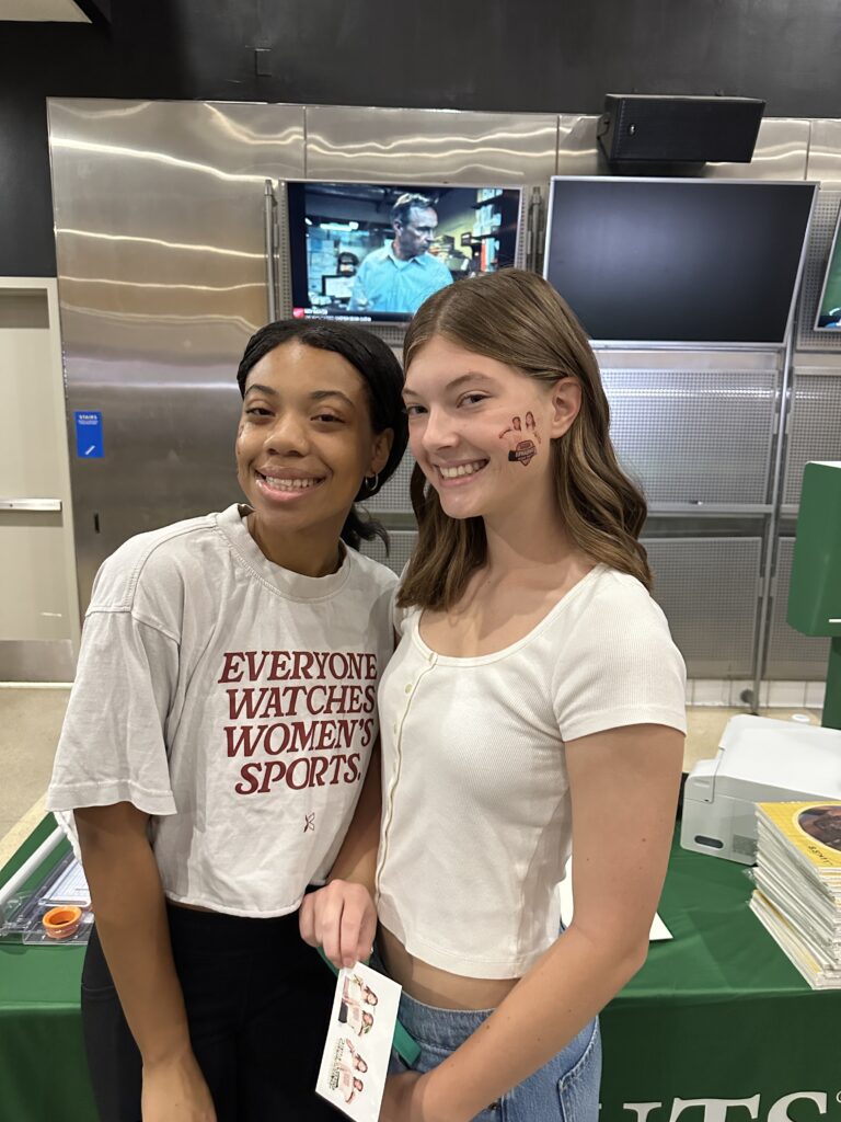 Two young women smile at an indoor event; one wears a "Everyone Watches Womens Sports" shirt, while the other sports painted paw prints—classic Shutterbox sports activations fun.