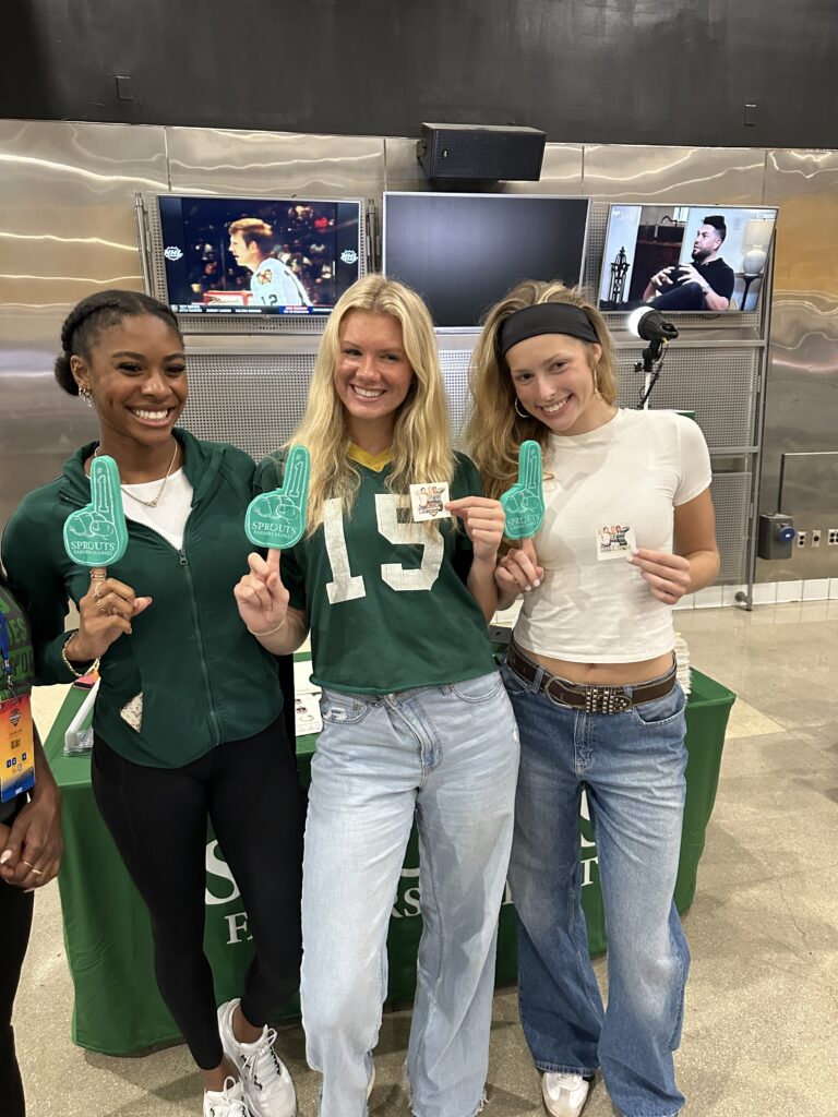 Three women pose indoors with green foam fingers and stickers, showcasing a creative fan engagement photo booth idea in front of a table draped with a green cloth; two TVs play behind them.