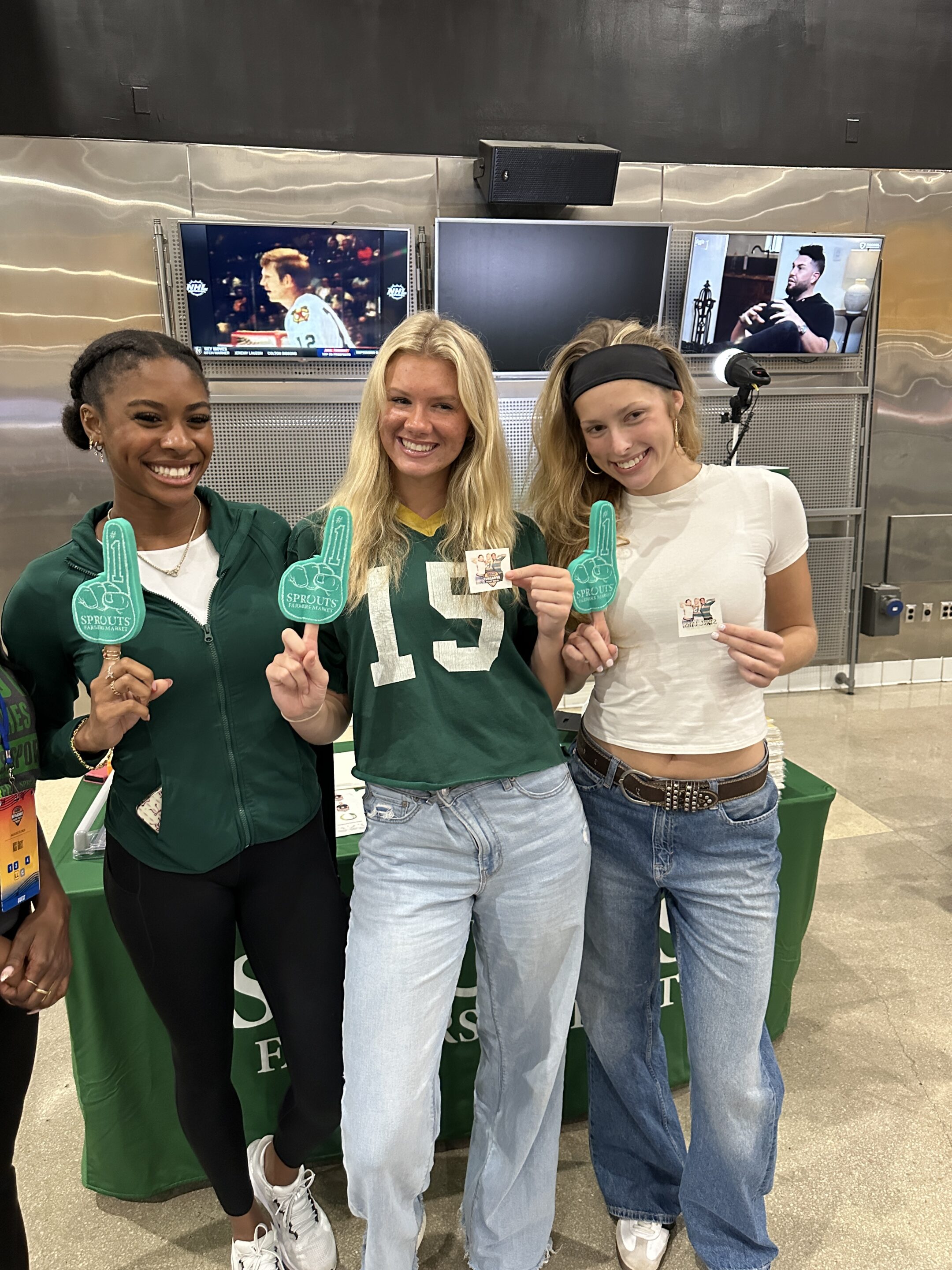 Three women pose indoors with green foam fingers and stickers, showcasing a creative fan engagement photo booth idea in front of a table draped with a green cloth; two TVs play behind them.