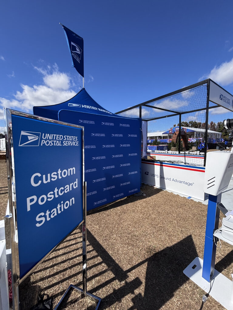 United States Postal Service booth with a Custom Postcard Station sign, tent, and equipment—plus an AI Photo Booth Las Vegas setup—arranged outdoors under a blue sky.