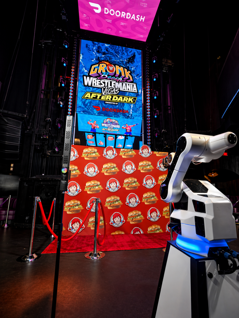 A robotic arm stands before a red step-and-repeat and a large screen at an Experiential marketing Las Vegas event, featuring Gronk WrestleMania Weekend After Dark with DoorDash and Wendy’s logos.