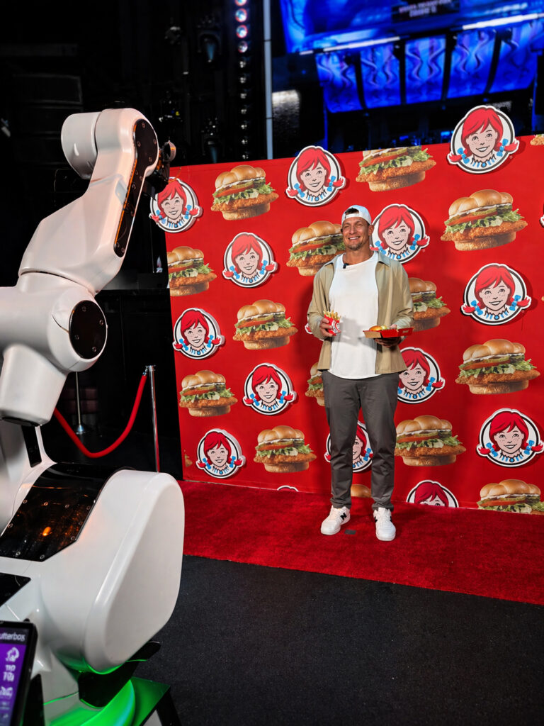 A man poses on a red carpet holding food and a drink in front of a Wendys backdrop, while a Glambot brand activation Las Vegas robotic arm faces him.