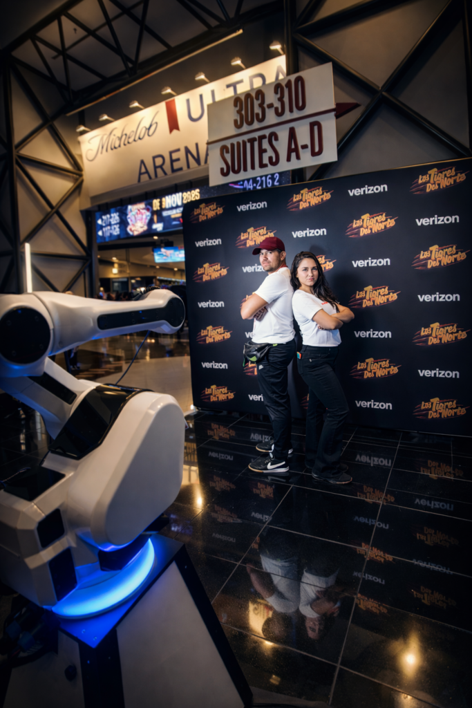 Two people pose back to back in front of a branded backdrop, a robotic arm in the foreground—perfect for custom photo booth for brand activations inside an arena concourse.