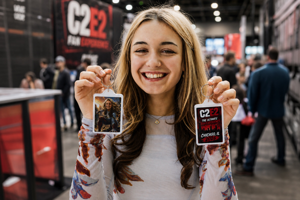 A smiling person holds two keychains at a convention; one highlights the Superhero AI Photo Booth Experience, the other displays C2E2 Fan Experience and event details.