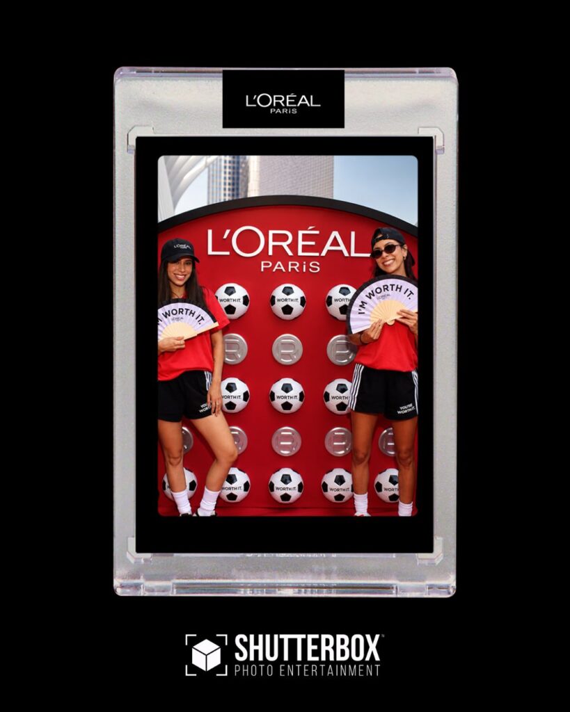 Two women in sportswear pose with soccer balls by a L'Oréal Paris display inside a Classic Photo Booth Rental Shutterbox entertainment frame.