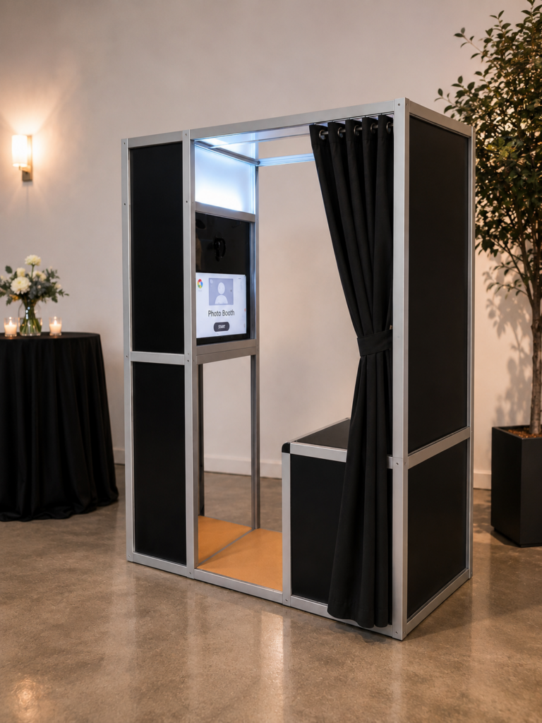 A modern Comic Con Photo Booth with black curtains stands indoors on a polished floor, near a round table with candles, flowers, and a potted plant in the background.