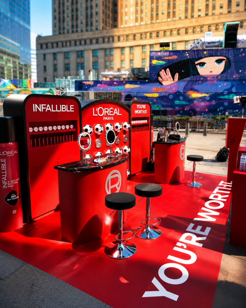 Red LOréal Paris outdoor booth with makeup displays, mirrors, stools on a branded mat—plus a Vintage Photo Booth Rental; city buildings and a mural in the background.