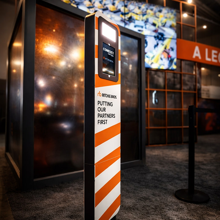 Freestanding kiosk with a digital screen displaying CONNECT at an indoor event, branded with Ritchie Bros and the slogan Putting our partners first.