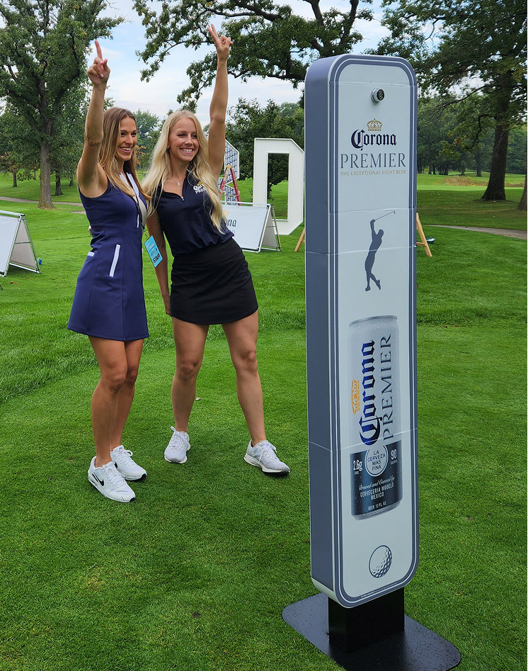 Two women in athletic outfits pose with arms raised next to a Corona Premier branded display on a grassy outdoor area, likely a golf course.