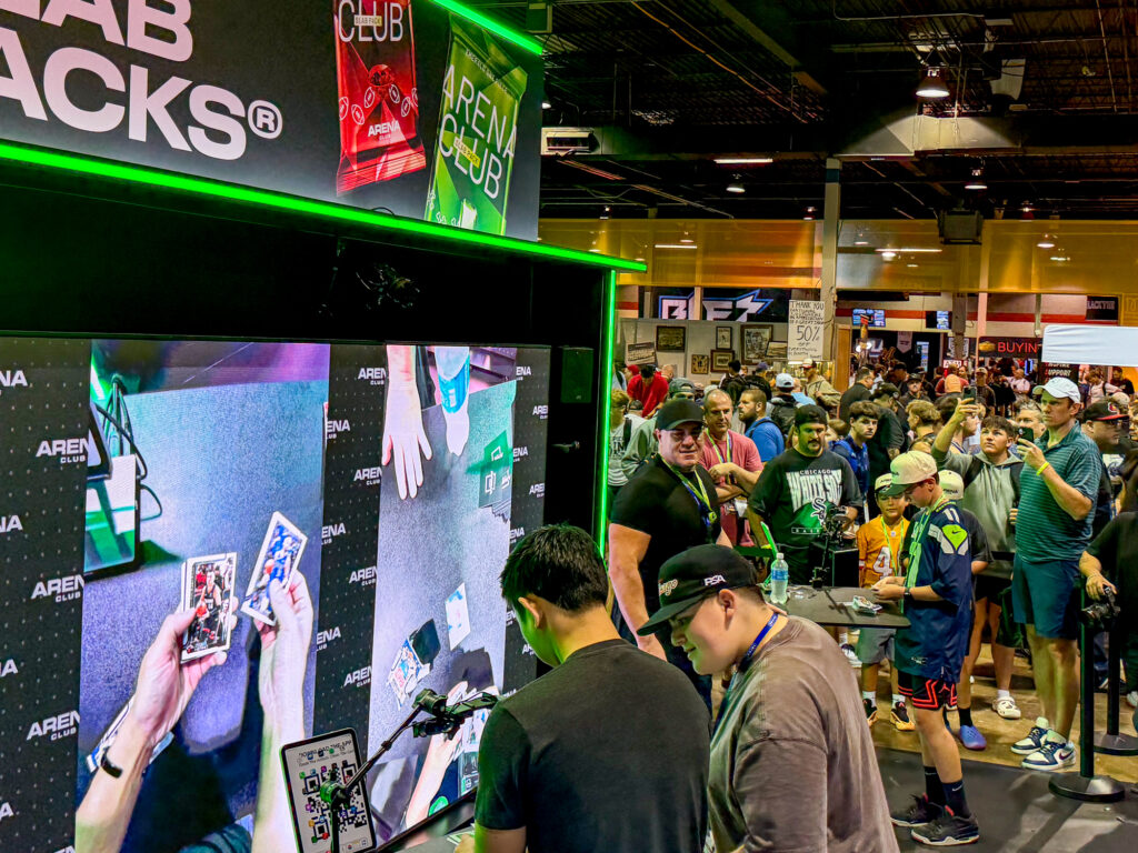 Two people open custom trading cards at a booth as a crowd watches, the action streamed on a large screen during an exciting sports marketing activation at the convention.