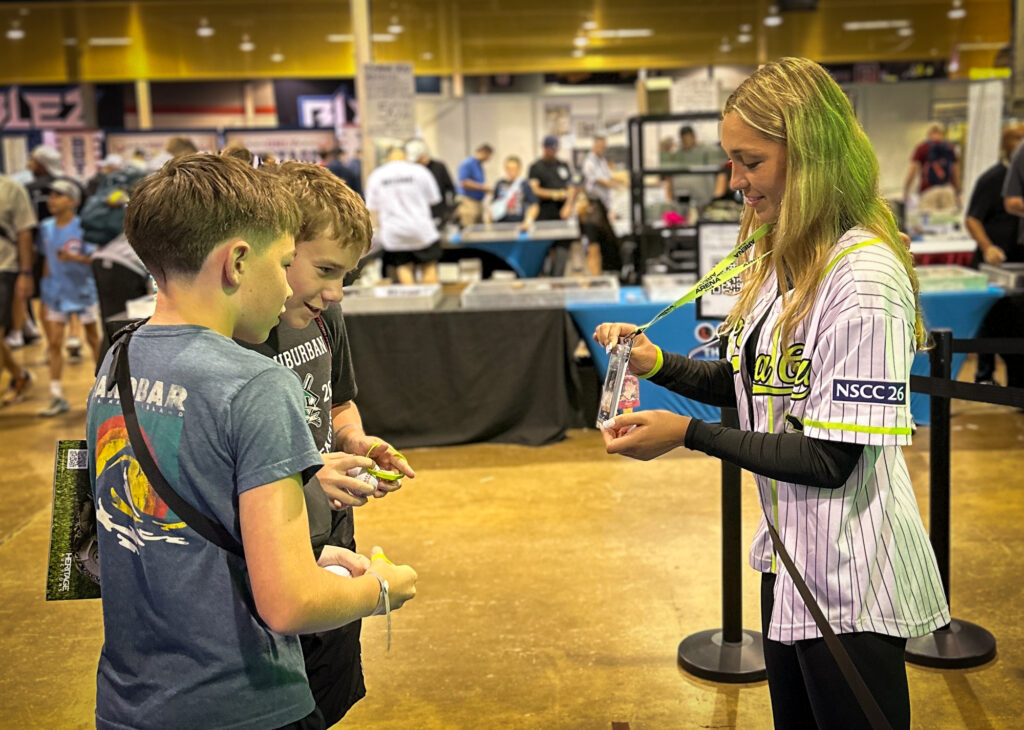 A woman shows sports trading cards to two boys at a convention, creating an engaging experiential marketing moment amid tables and attendees in the background.