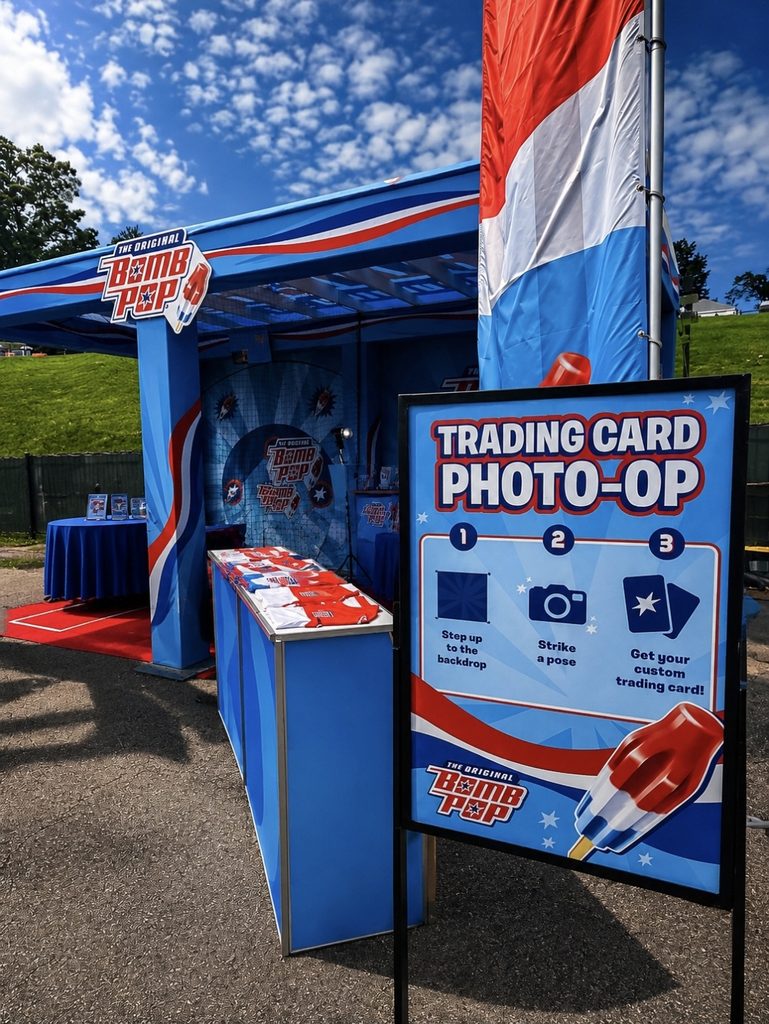 A Bomb Pop-branded booth at The National Sports Collectors Convention offers a Trading Card Photo Booth Rental and photo-op instructions under a partly cloudy sky in an outdoor setting.